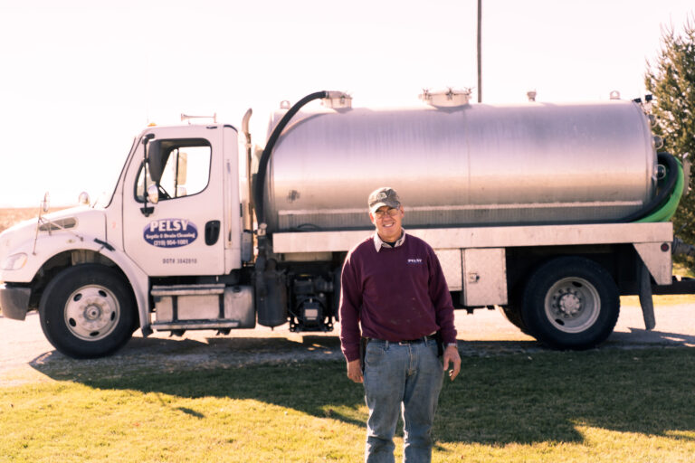 man standing in front of a pelsy septic pumper truck with the sun in the background